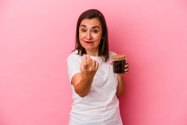 Keski-ikäinen valkoihoinen nainen pitää kahvipurkkia eristettynä vaaleanpunaisella taustalla osoittaen sormella sinua ikään kuin kutsuva tulla lähemmäksi. - Valokuva, kuva