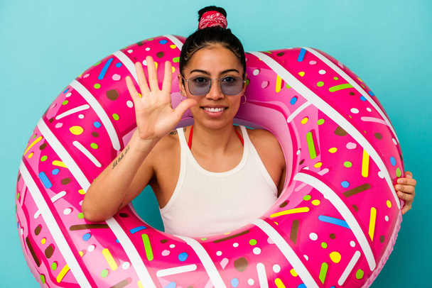 Jovem mulher latina segurando um donut inflável isolado no fundo azul sorrindo alegre mostrando número cinco com os dedos. - Foto, Imagem