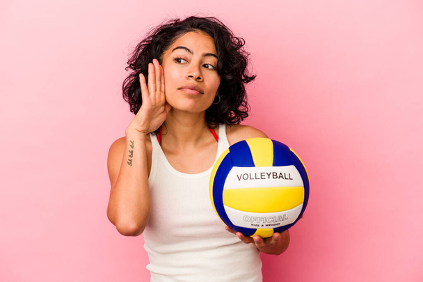 Joven latina sosteniendo una pelota de voleibol aislada sobre fondo rosa tratando de escuchar un chisme. - Foto, imagen