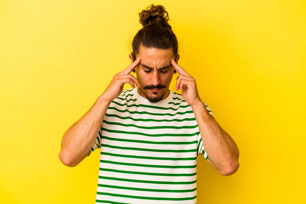 Junge kaukasische Mann mit langen Haaren isoliert auf gelbem Hintergrund berührt Schläfen und hat Kopfschmerzen. - Foto, Bild