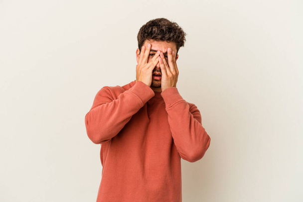Young caucasian man isolated on white background blink at the camera through fingers, embarrassed covering face. - Фото, изображение