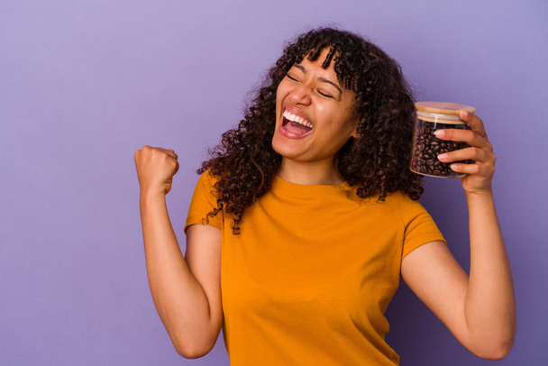 Молода змішана расова жінка тримає пляшку кавових зерен ізольовано на фіолетовому фоні, піднімаючи кулак після перемоги, концепція переможця
. - Фото, зображення
