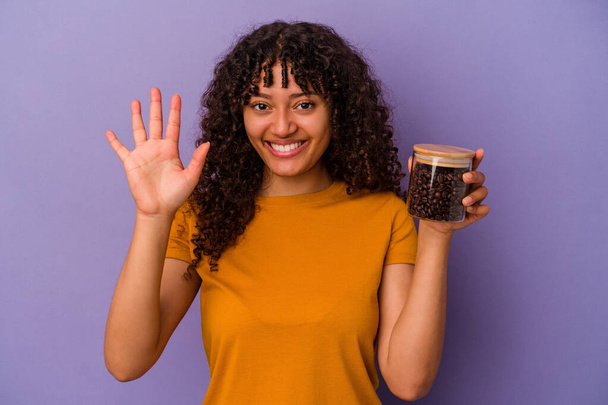 Nuori sekarotuinen nainen pitelee kahvipapuja pullo eristetty violetti tausta hymyilevä iloinen osoittaa numero viisi sormin. - Valokuva, kuva