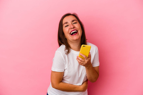 Middle age caucasian woman holding mobile phone isolated on pink background laughing and having fun. - 写真・画像