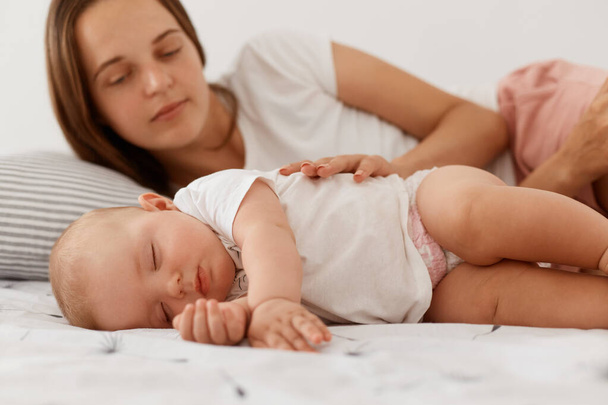 Young adult female with dark hair lying with baby in bed, looking at daughter to sees sleeping she or not, woman wearing white casual t shirt, happy motherhood. - Fotó, kép