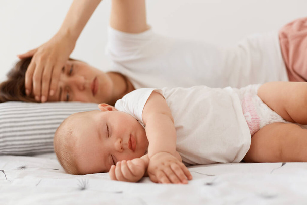 Tired exhausted female wearing white casual style t shirt lying near sleeping baby, keeping palm on forehead, lying with child, having sleepless night, motherhood. - Photo, image