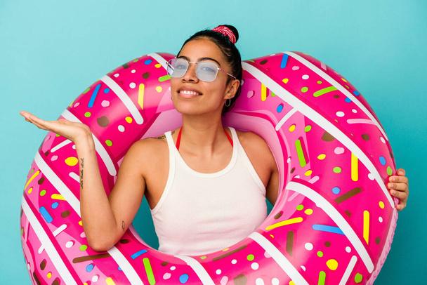 Young latin woman holding an inflatable donut isolated on blue background showing a copy space on a palm and holding another hand on waist. - Fotoğraf, Görsel