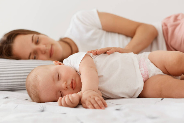 Indoor shot of smiling female lying with sleeping girl daughter, resting together with her lovely baby, woman wearing white casual t shirt looking at infant girl with positive emotions. - Fotó, kép