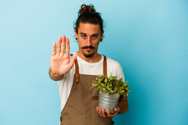 Νεαρός κηπουρός καυκάσιος άνδρας που κρατά ένα φυτό απομονωμένο σε μπλε φόντο στέκεται με τεντωμένο χέρι που δείχνει στοπ, εμποδίζοντάς σας. - Φωτογραφία, εικόνα