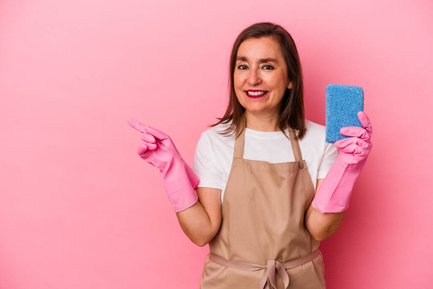 Mujer caucásica de mediana edad limpiando casa aislada sobre fondo rosa sonriendo y señalando a un lado, mostrando algo en espacio en blanco. - Foto, imagen