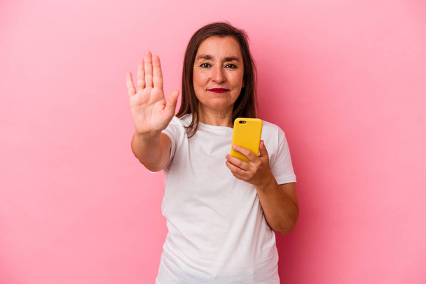 Mujer caucásica de mediana edad sosteniendo teléfono móvil aislado sobre fondo rosa de pie con la mano extendida que muestra señal de stop, lo que le impide. - Foto, Imagen