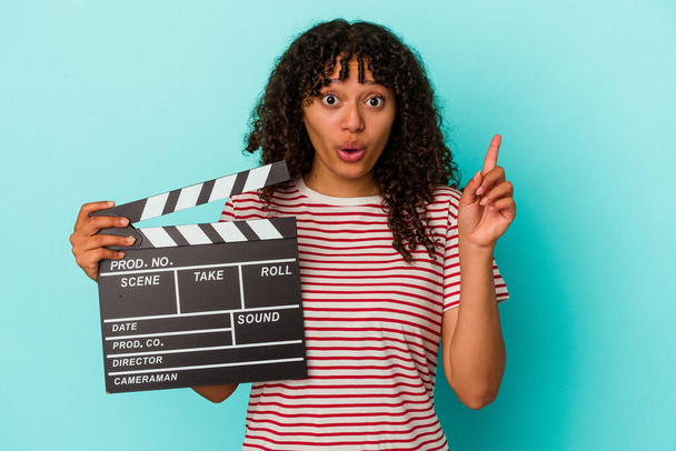 Jonge gemengde ras vrouw met een clapperboard geïsoleerd op blauwe achtergrond met een geweldig idee, concept van creativiteit. - Foto, afbeelding