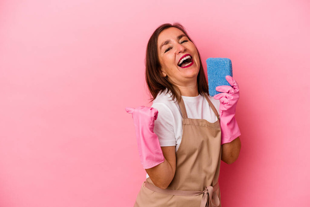 Middle age caucasian woman cleaning home isolated on pink background points with thumb finger away, laughing and carefree. - Фото, изображение