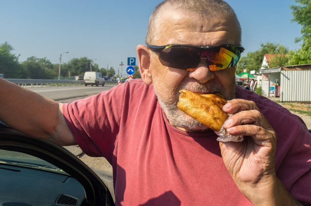 Portarait of hungry Cacasiam senior driver wearing sunglasses eating patty near his car standing on Ukrainian road - Photo, Image
