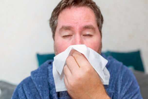 opieka zdrowotna, ludzie i medycyna - chory człowiek dmuchający nos w domu. chory mężczyzna w domu ma problemy ze zdrowiem, objawy grypy lub gorączki, zmęczony facet cierpi na chorobę kichania trzymając tkankę. - Zdjęcie, obraz