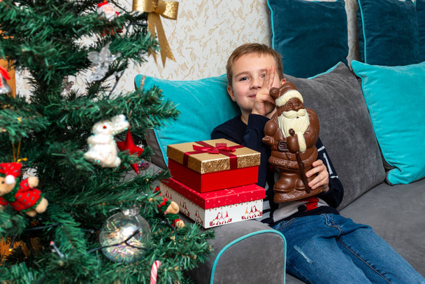 Fiú ül a kanapén kezében csokoládé Mikulás karácsonyi ajándékok.Mellette a karácsonyfa díszített otthon. - Fotó, kép