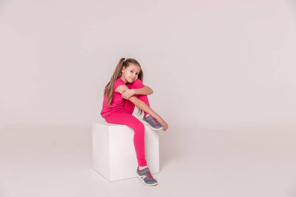 beautiful sports girl in a pink T-shirt and pants on a white isolated background. child sitting on a white cube. girl with tails. Copy space - Foto, Imagem