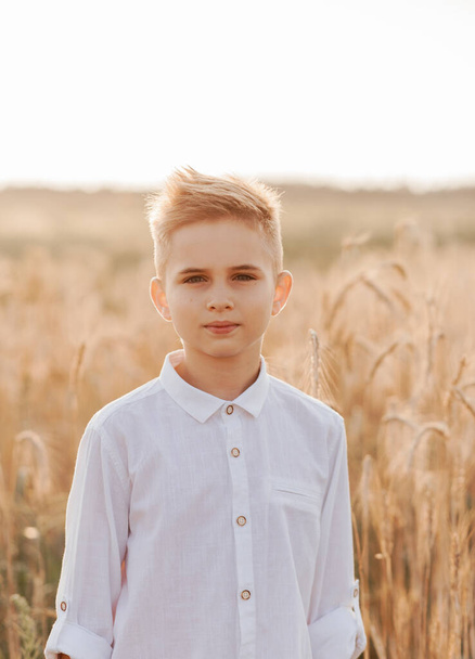 A small handsome boy blonde in a white shirt and white pants in nature in the summer. Happy childhood. Positive emotions - Photo, Image
