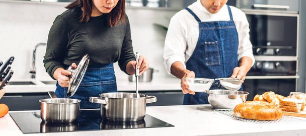 Young asian family couple having fun standing near stove and cooking together.Happy couple looking and smelling tasting fresh delicious from soup in a pot with steam at white interior kitchen - Foto, Imagem