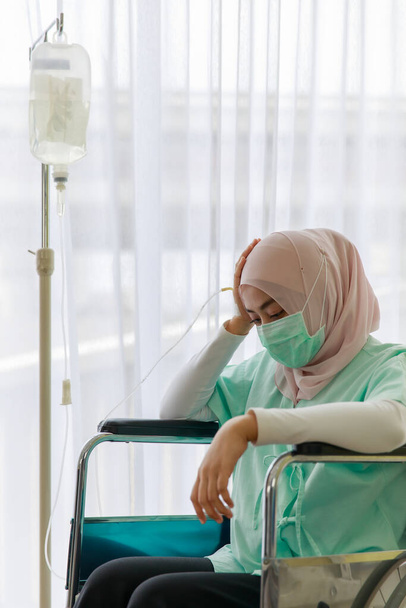 Portrait of ill Muslim wearing patient gown and face mask sitting on a wheelchair with saline fluid bag attach in hospital. Covid 19 pandemic concept. - Photo, Image