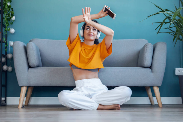 Shot of motivated young woman listening to music with smartphone while dancing sitting on the floor at home. - Foto, Bild