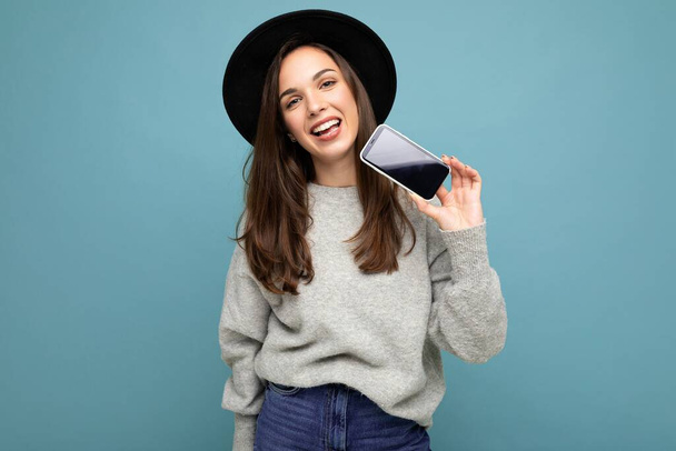 Досить молода усміхнена жінка в чорному капелюсі і сірий светр тримає телефон, дивлячись на камеру ізольовано на фоні
 - Фото, зображення