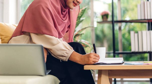 Genç, gülümseyen Müslüman bir kadın dizüstü bilgisayarını kullanarak rahatlıyor ve evde video konferansı yapıyor. Ev konseptinden çalışma. - Fotoğraf, Görsel