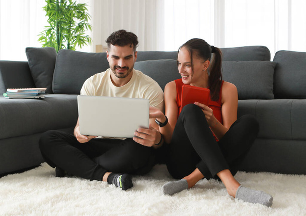 Szczęśliwa atrakcyjna młoda kaukaska para siedzieć na białym dywanie i korzystać z laptopa do rozrywki online i zakupów w salonie. Aktywność relaksacyjna w domu podczas koncepcji blokady - Zdjęcie, obraz