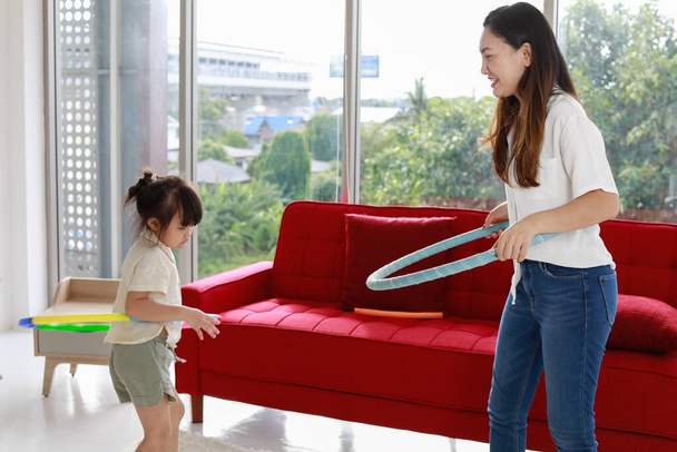 Asian beautiful young long black hair single mom stand smile playing hula hoop together with little cute daughter girl next to red sofa and bed in living bedroom with clear glass windows background. - Fotó, kép