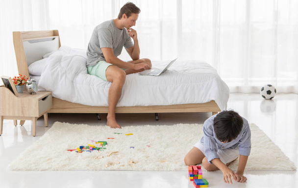 Otec sedí na posteli a pracuje s notebookem, zatímco jeho smíšený syn si hraje na barevný dřevěný blok v domácí ložnici. Nápad na životní styl práce z domova - Fotografie, Obrázek