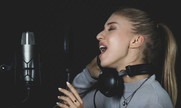 Молодая певица исполняет песню возле микрофона в профессиональной студии звукозаписи со звуконепроницаемой стеной - Фото, изображение