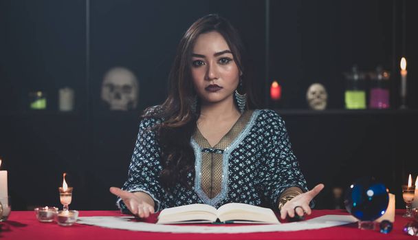 Genç ve sevimli Asyalı kadın falcı çingene tarzı giysiler içinde elinde kitapla loş odada fal bakarken kameraya bakıyor. - Fotoğraf, Görsel