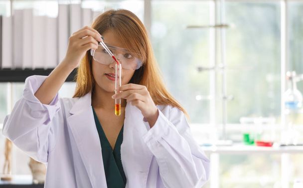 Científica asiática joven y exitosa en bata de laboratorio blanca y gafas de seguridad vertiendo solución de reactivo de color de Erlenmeyer frasco en vaso de precipitados de vidrio en laboratorio lleno de equipo científico y libros. - Foto, Imagen