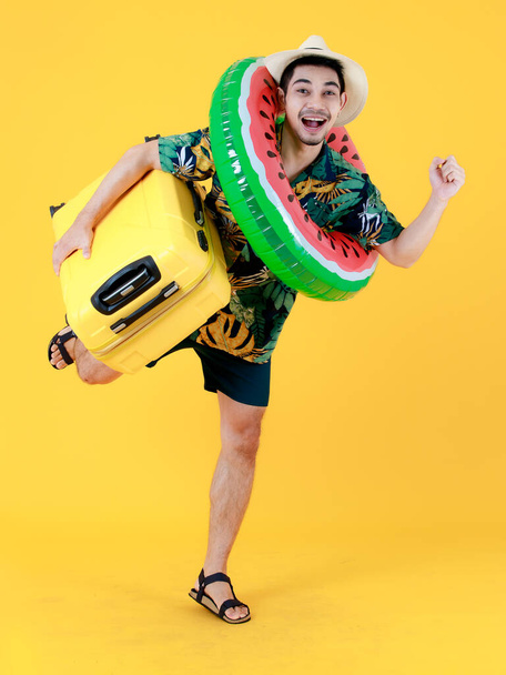Joyeux jeune homme asiatique en chemise hawaïenne colorée se tient sur une jambe tient une valise jaune et anneau de natation. Portrait studio plein corps drôle sur fond jaune. Concept de voyage vacances d'été - Photo, image