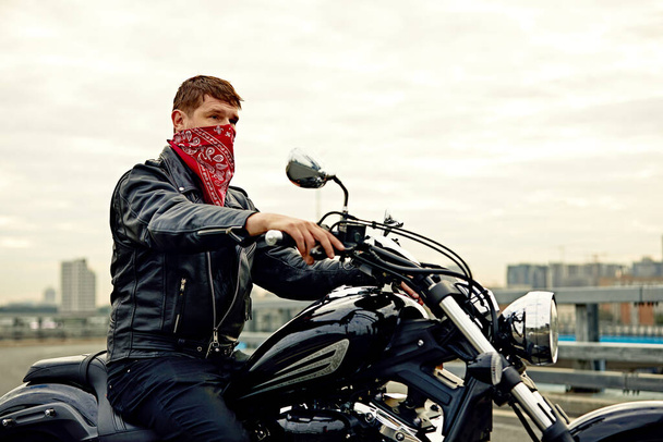 Portrait d'un homme élégant vêtu d'une veste en cuir noir et d'un bandana assis sur sa moto rétro sur mesure, regardant ailleurs, bâtiments de la grande ville en arrière-plan. - Photo, image