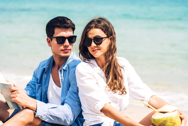 Νεαρό ζευγάρι πίνει χυμό καρύδας και διαβάζει ένα βιβλίο για την τροπική παραλία.Ζευγάρι χαλαρώνει στην παραλία.Διακοπές - Φωτογραφία, εικόνα
