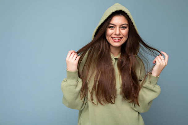Портрет привлекательной молодой кавказской улыбающейся брюнетки-модели в модной хаки-толстовке с капюшоном на синем фоне с копировальным пространством. Позитивная концепция - Фото, изображение