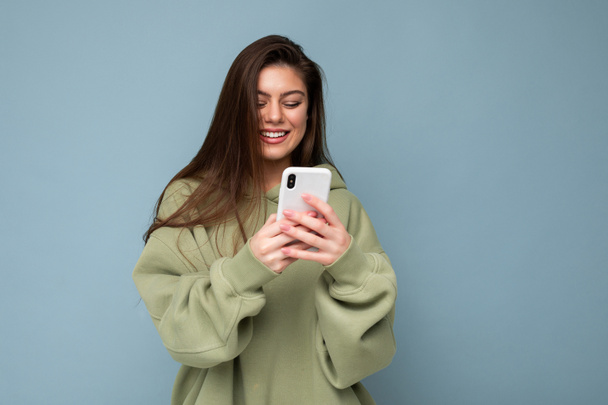 Sorridente Bella giovane donna bruna che indossa elegante felpa verde con cappuccio utilizzando sms di scrittura del telefono cellulare isolati sullo sfondo guardando lo schermo dello smartphone divertirsi - Foto, immagini
