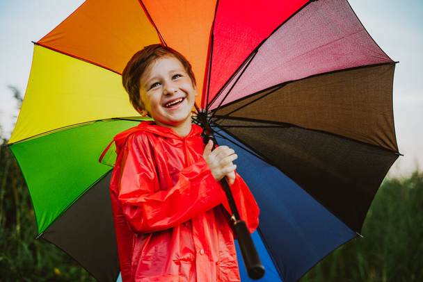 Portrét usměvavého školáka s duhovým deštníkem v parku. Dítě drží na rameni barevný deštník. Veselé dítě v červeném plášti, držící venku vícebarevný deštník - Fotografie, Obrázek
