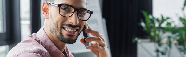 Hymyilevä muslimi liikemies silmälaseissa puhuu älypuhelimella toimistossa, banneri  - Valokuva, kuva