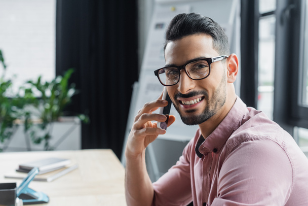 Аравійський бізнесмен у окулярах посміхається перед камерою, розмовляючи на смартфоні в офісі.  - Фото, зображення