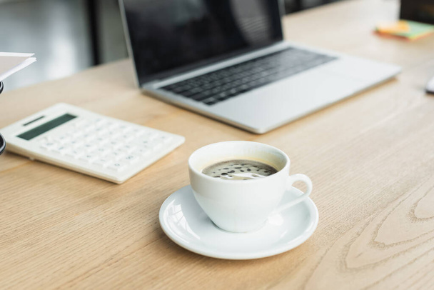 Cup of coffee near blurred laptop and calculator in office  - Φωτογραφία, εικόνα