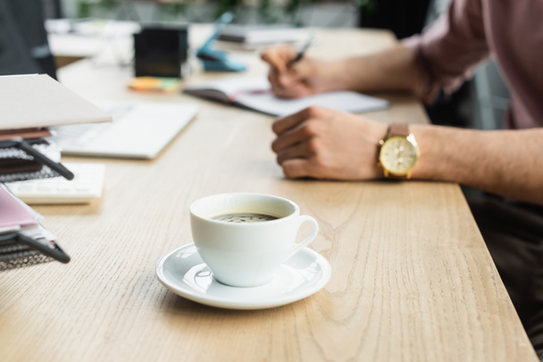 Обрезанный вид на чашку кофе на столе возле размытого бизнесмена в офисе  - Фото, изображение