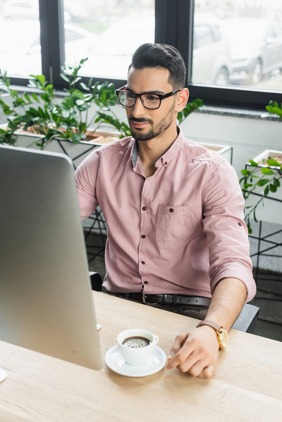 Muslim businessman in eyeglasses using computer near cup of coffee in office  - Zdjęcie, obraz
