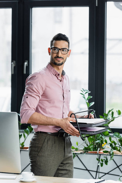 Arabský podnikatel drží tác s papíry v blízkosti počítače a šálek kávy v kanceláři  - Fotografie, Obrázek