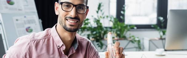 Allegro uomo d'affari musulmano in occhiali che tiene il disinfettante per le mani in ufficio, striscione  - Foto, immagini