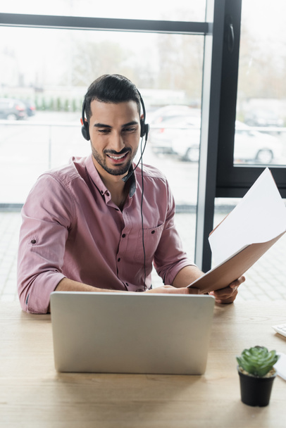 Pozitív muszlim üzletember fülhallgató papír mappában videohívás közben az irodában  - Fotó, kép