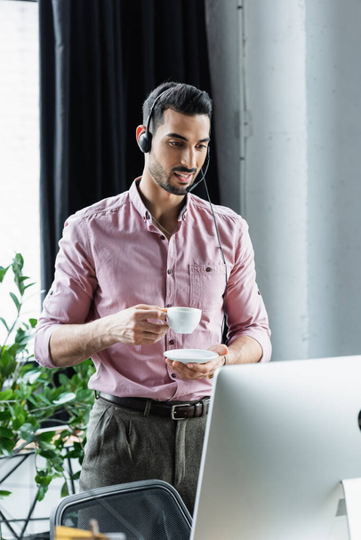 オフィスでコンピュータの近くにコーヒーのカップを保持ヘッドセットで笑みを浮かべてイスラム教徒のビジネスマン  - 写真・画像