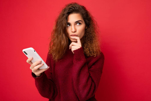 Fotografie hezké přemýšlivé mladé ženy s kudrnaté vlasy na sobě tmavě červený svetr izolované na červeném pozadí držení mobilního telefonu při pohledu na stranu a přemýšlení - Fotografie, Obrázek