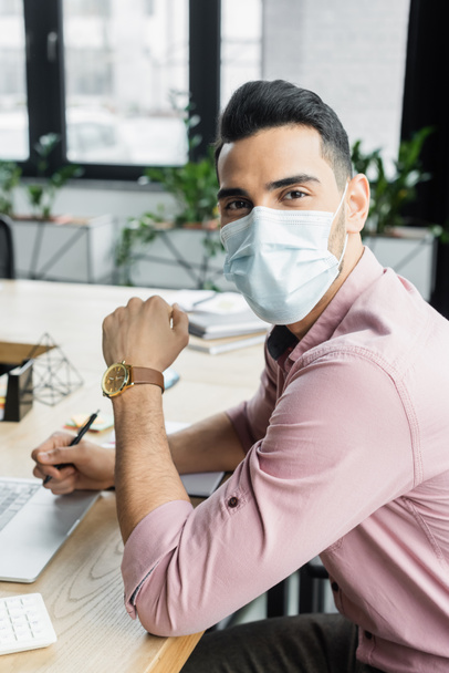 Arabský podnikatel v lékařské masce při pohledu do kamery v blízkosti rozmazaného notebooku v kanceláři  - Fotografie, Obrázek